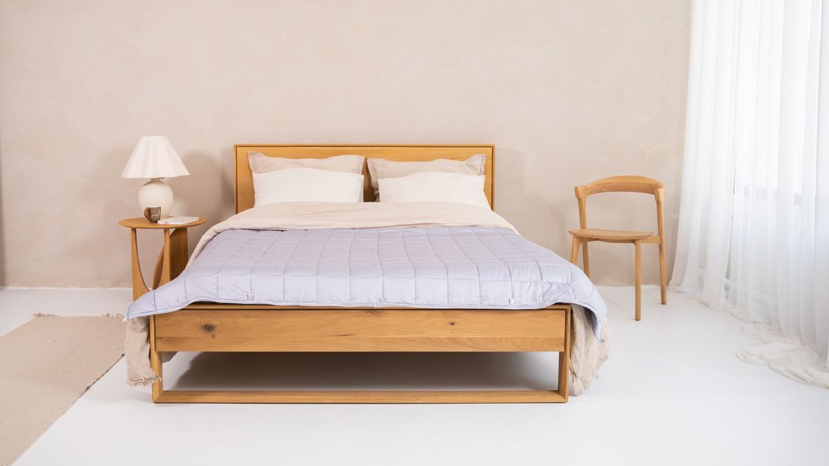 A solid oak bed frame with a low headboard in a pale, sunlit bedroom