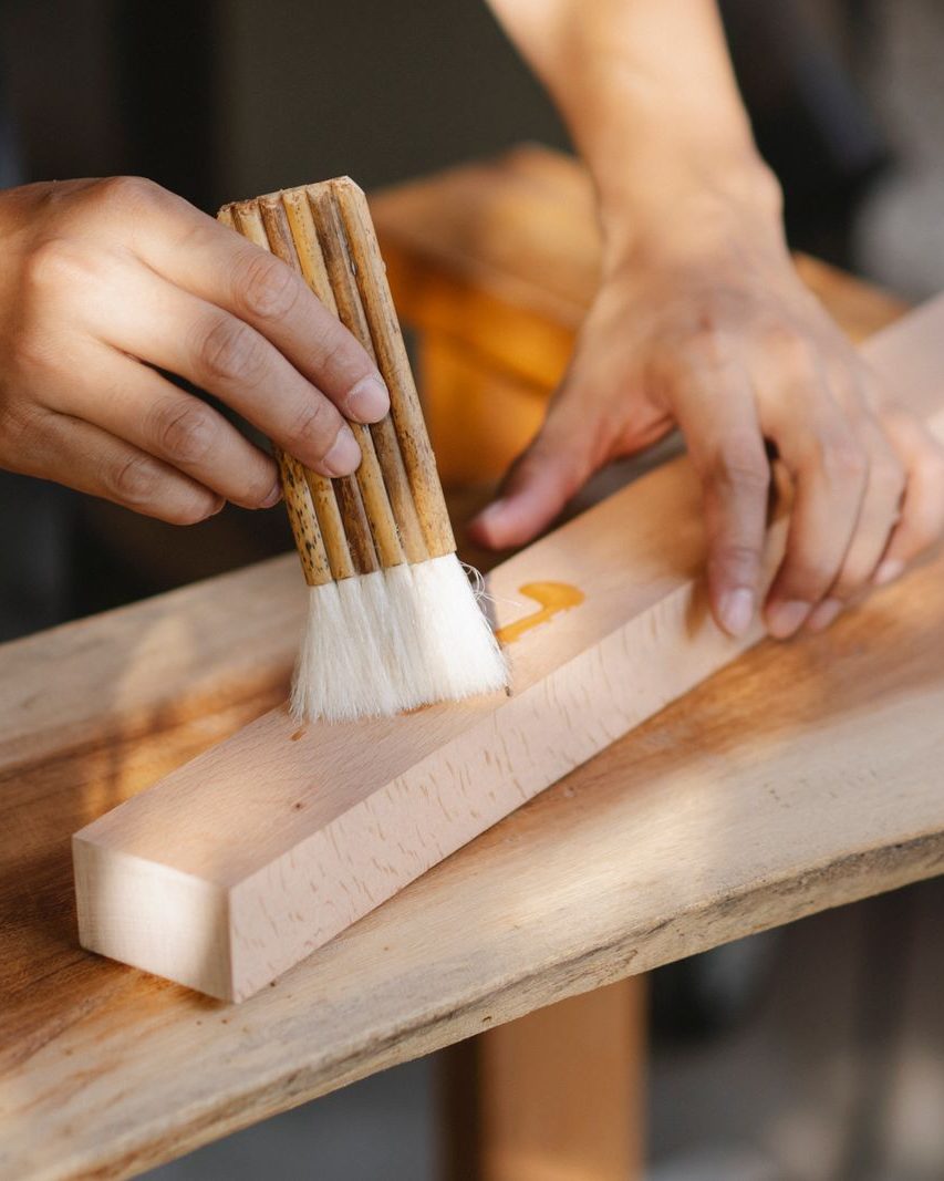 Hands brushing oil into a plank of pale wood on a workbench